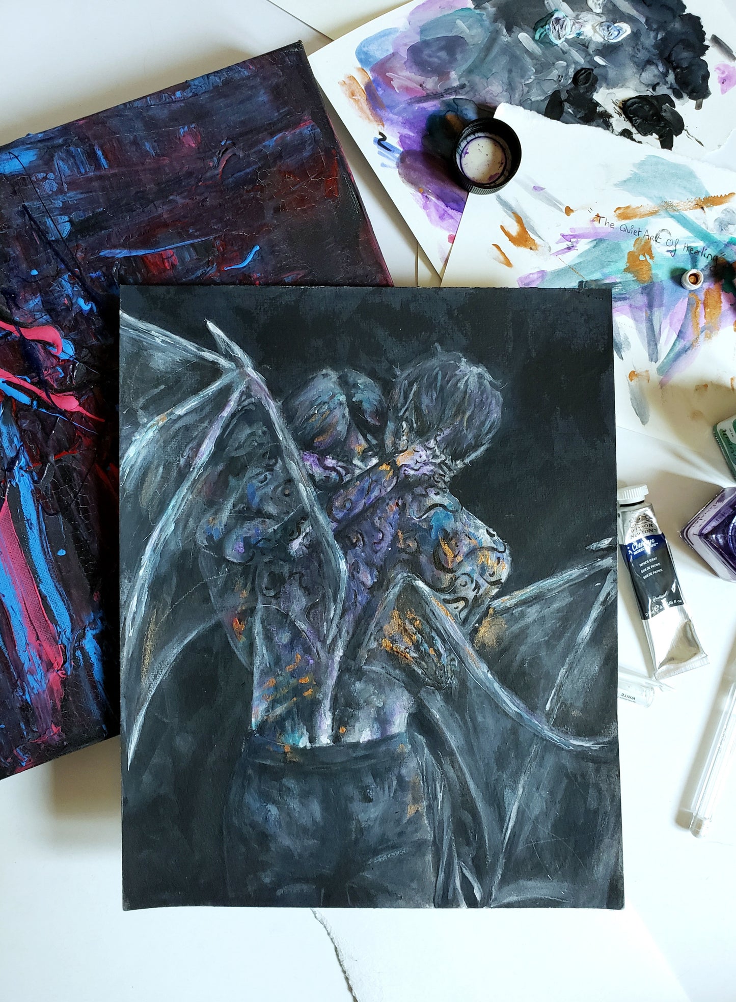 Back view of a Fantasy fae couple embracing each other and covered in paint. Grey and black nuances with hints of purple, orange, blue colors on the male's back. 